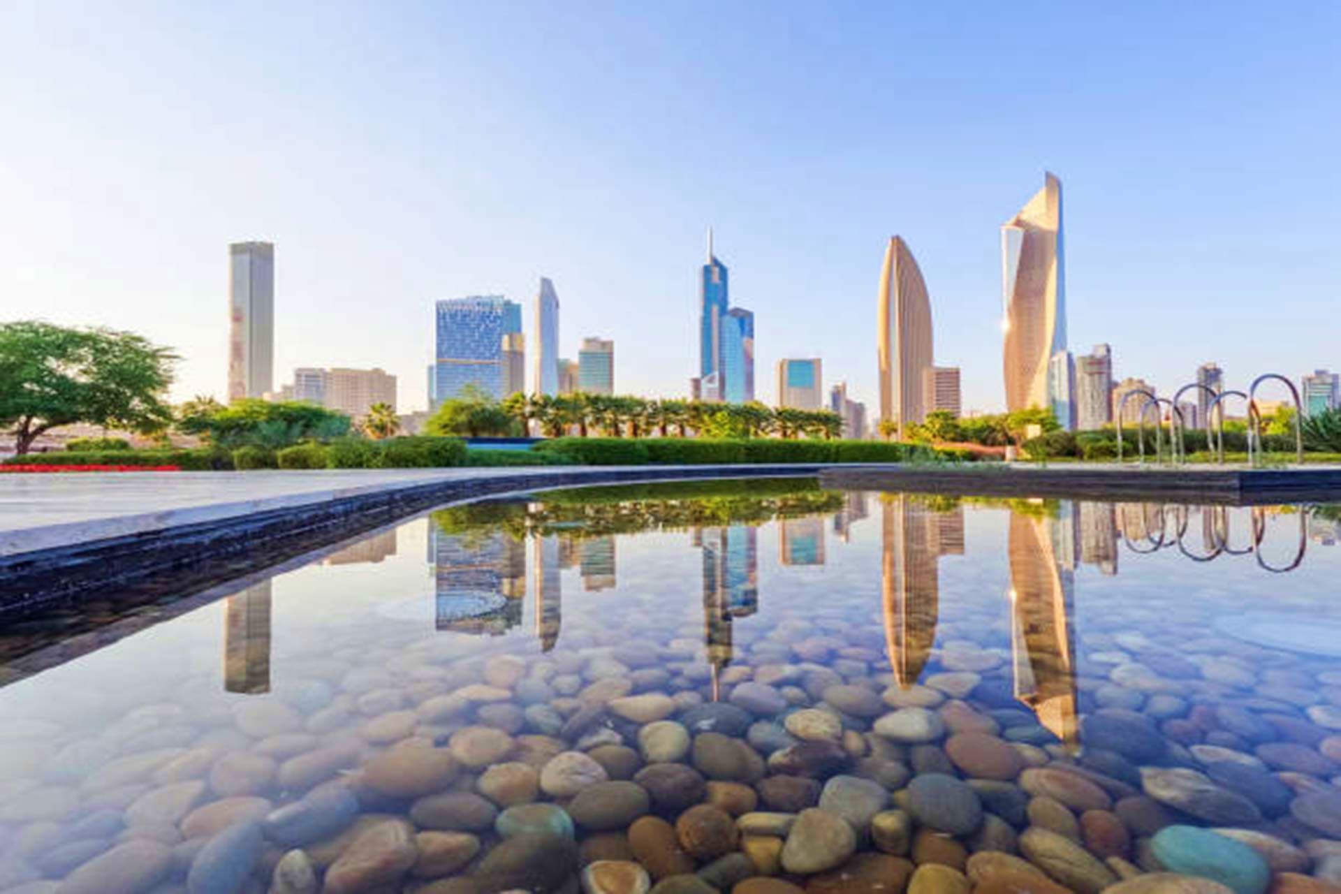 Kuwait Visa Kuwait City skyline with modern skyscrapers reflected in waterfront pool, palm trees and landscaping visible